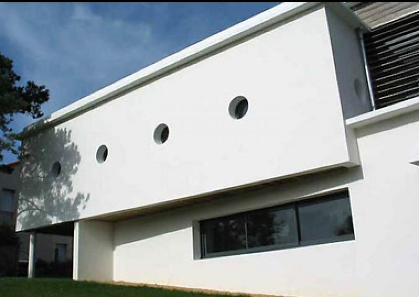 Façade d'une maison contemporaine enduite de blanc, percée de petits hublots circulaires et d'une longue baie vitrée, sous un ciel bleu.