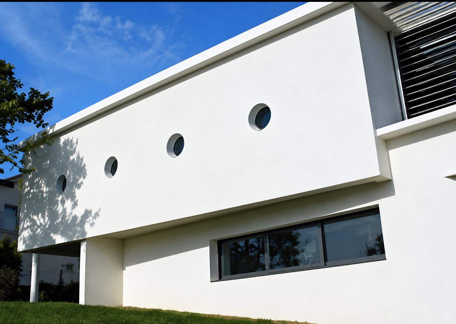 Façade d'un bâtiment moderne blanc percée de petites fenêtres rondes et d'une longue baie vitrée, sous un ciel bleu.