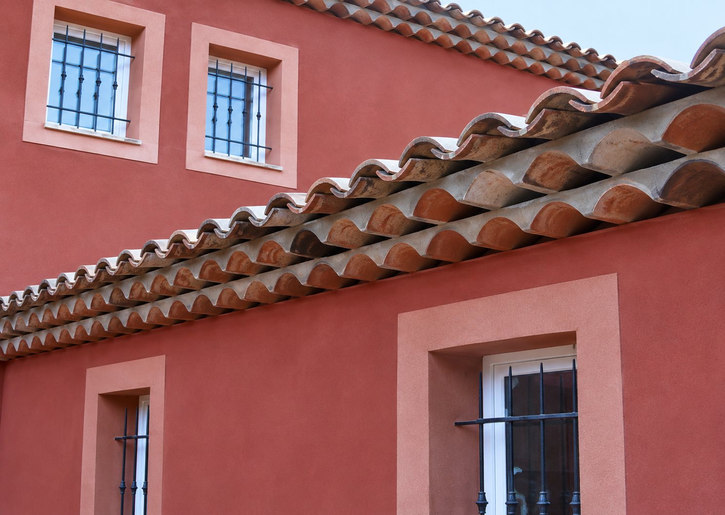 Façade d'une maison rouge aux fenêtres grillagées, surmontée d'un toit de tuiles canal en terre cuite sous un ciel bleu.