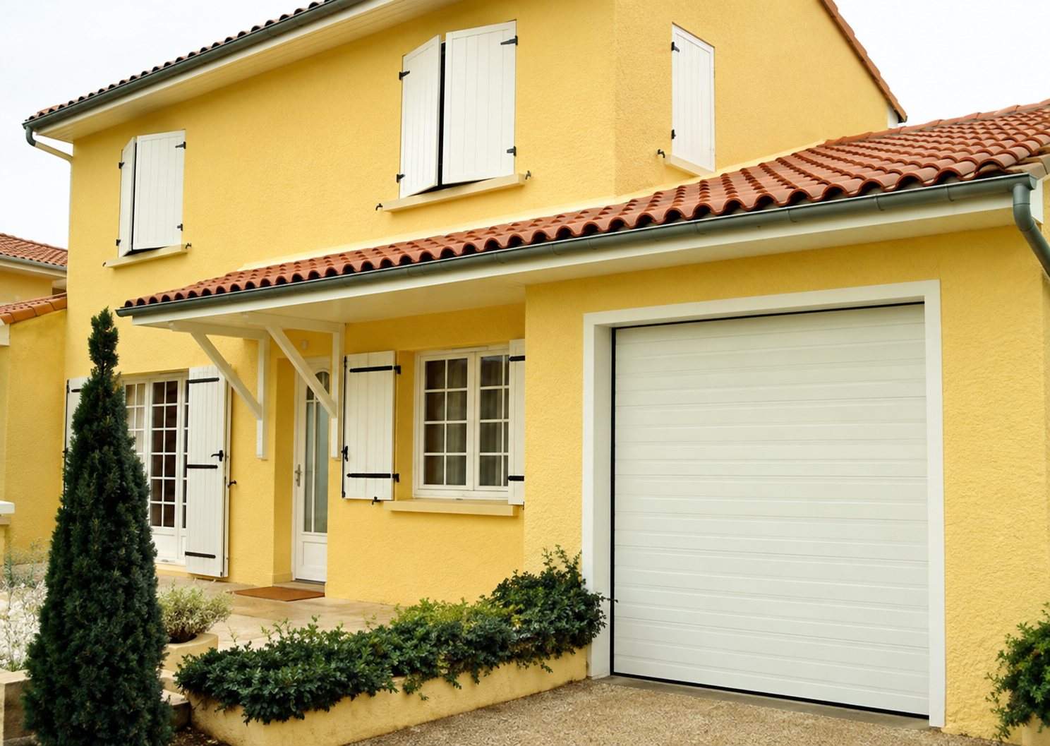 Façade d'une maison jaune avec volets blancs, une porte de garage blanche, un toit en tuiles et un petit conifère devant.