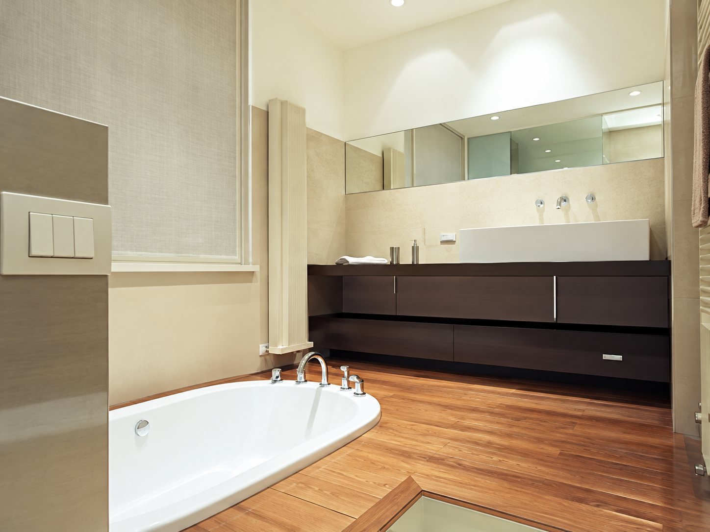 Salle de bain contemporaine avec une baignoire blanche encastrée, un sol en bois et un meuble vasque sombre sous un grand miroir.