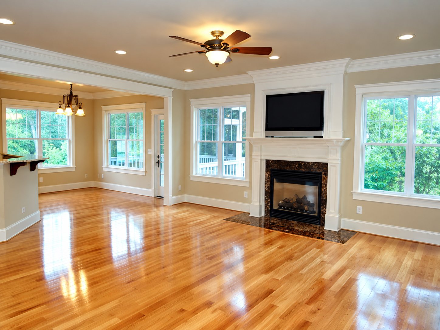 Grande pièce vide et lumineuse avec un parquet en bois verni, une cheminée surmontée d'un écran, un ventilateur de plafond et de larges fenêtres.