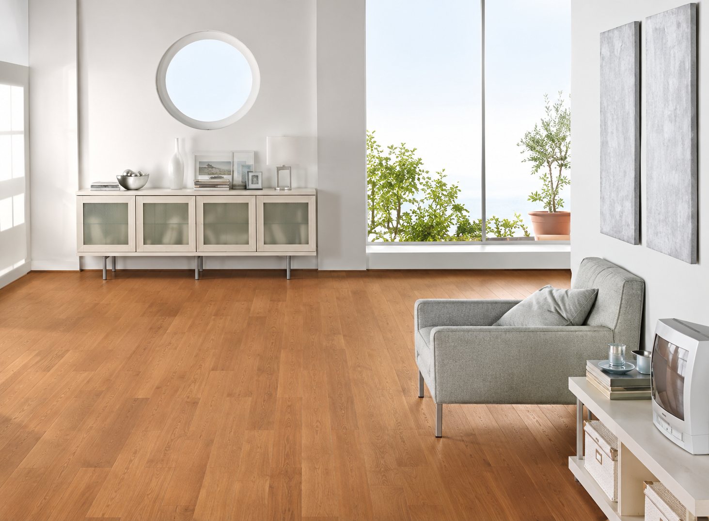 Un salon lumineux au parquet en bois clair, avec un buffet blanc, un miroir rond, un fauteuil gris et de grandes fenêtres.