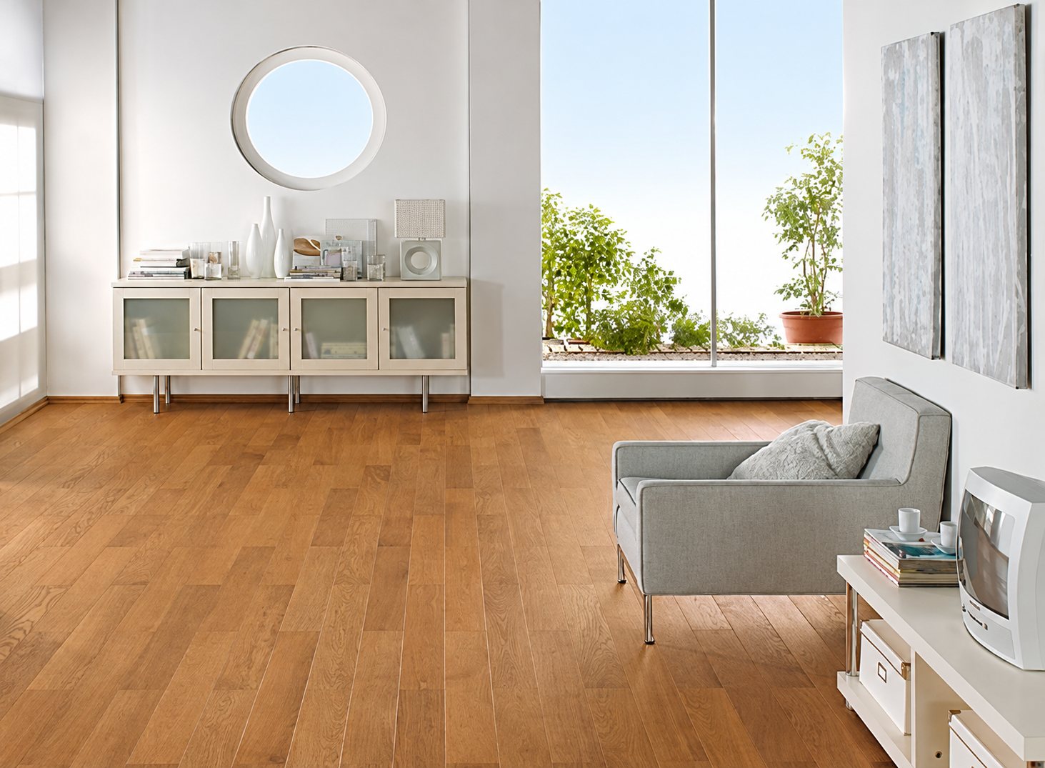 Un salon moderne et lumineux avec un parquet en chêne miel, un buffet blanc à portes vitrées, un miroir rond, un fauteuil gris et une grande baie vitrée.
