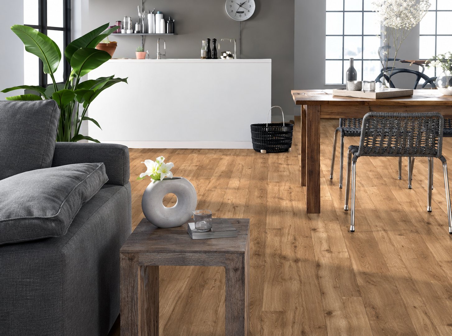 Un salon lumineux avec un sol en parquet clair, un canapé gris, une table basse en bois et une table à manger au fond.