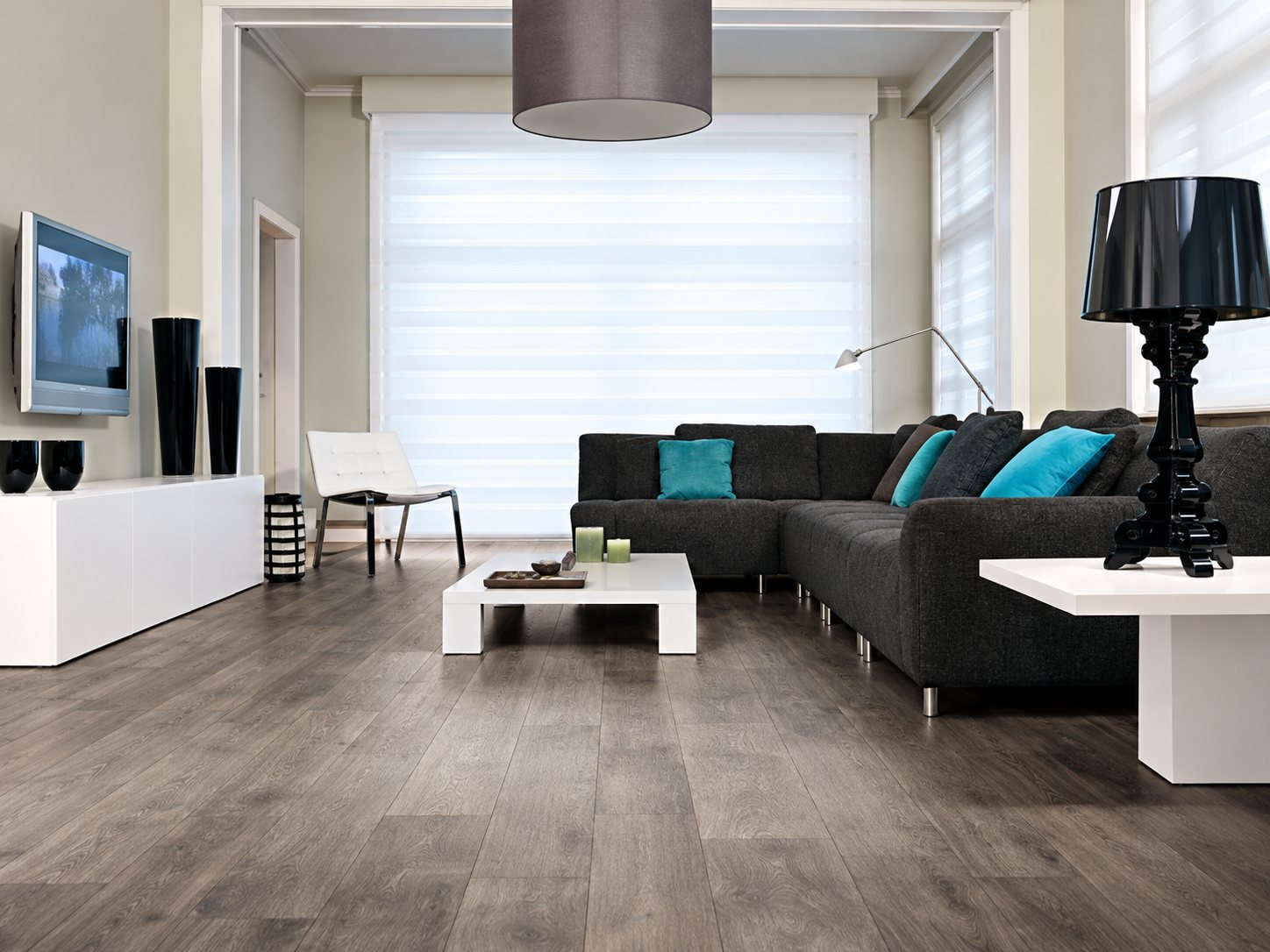 Un salon lumineux au parquet gris comprend un canapé d'angle sombre avec des coussins turquoise, une table basse blanche et une lampe noire.