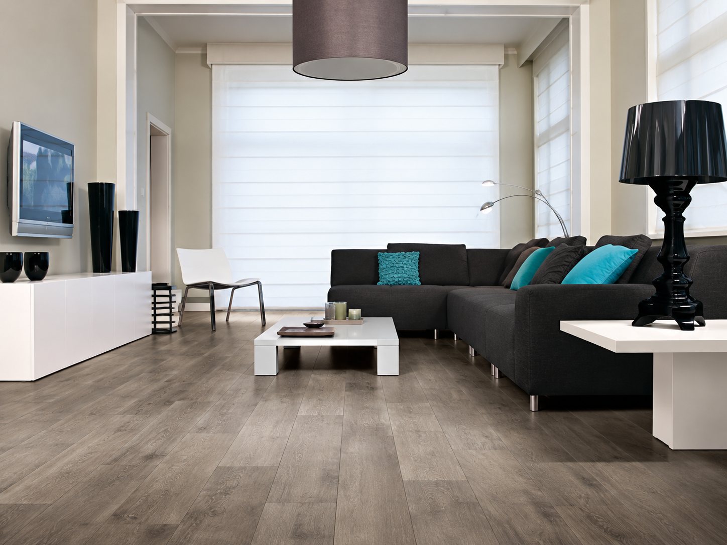 Salon lumineux au sol en parquet gris, avec un canapé d'angle sombre, des coussins turquoise, une table basse blanche et une lampe noire.