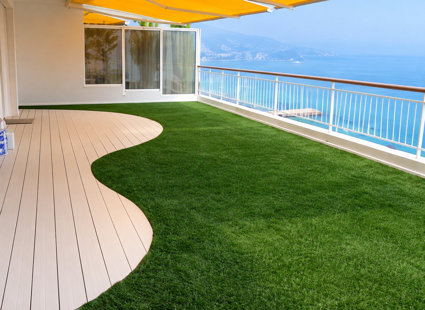 Une terrasse en bord de mer aménagée avec un gazon synthétique vert aux contours courbes, bordé d'un platelage clair et d'une rambarde en verre.